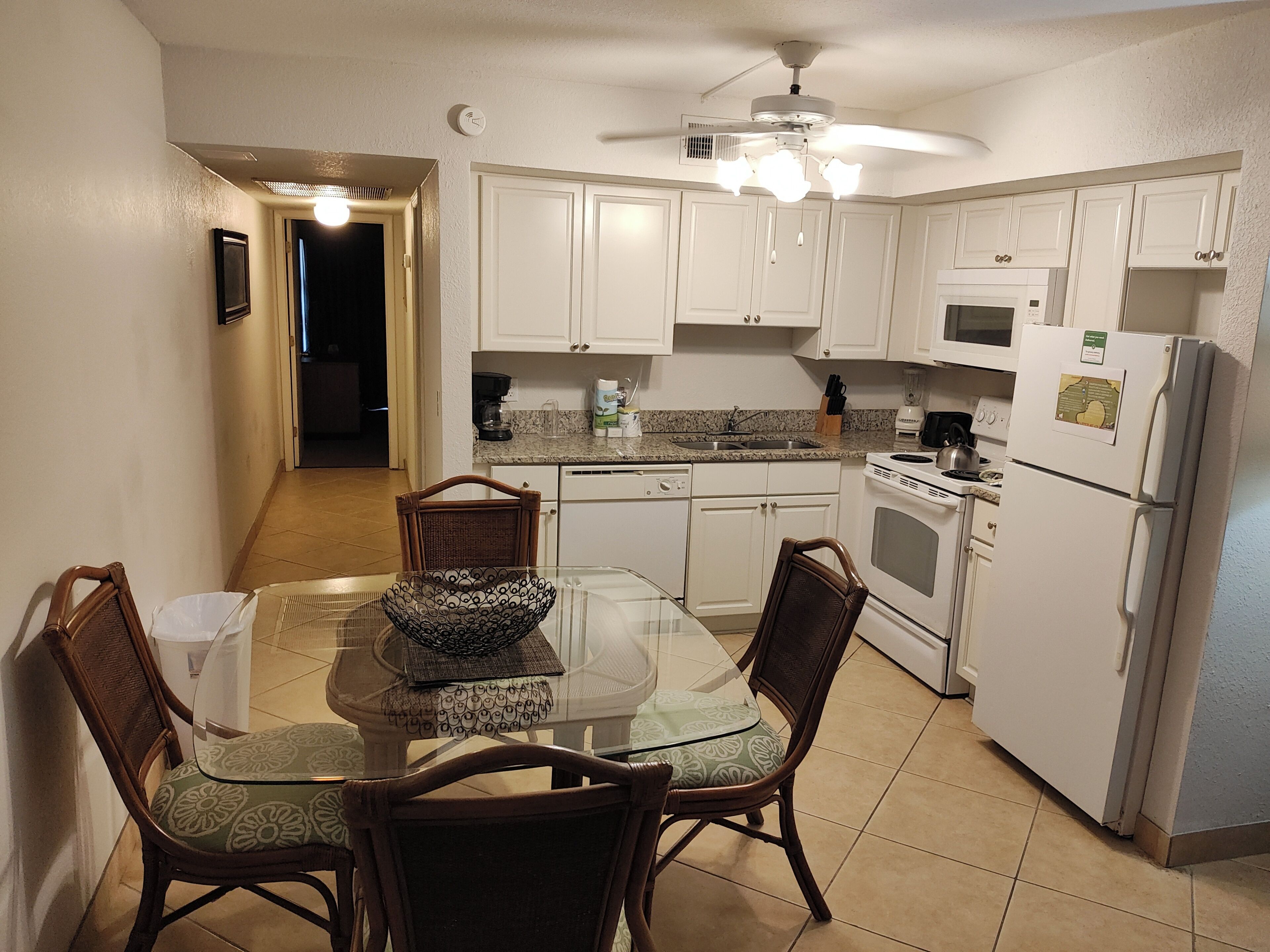 Una cocina compacta con armarios blancos, una mesa de comedor con sillas, un ventilador de techo y un refrigerador.