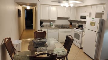 A compact kitchen with white cabinets, a dining table with chairs, a ceiling fan, and a refrigerator.