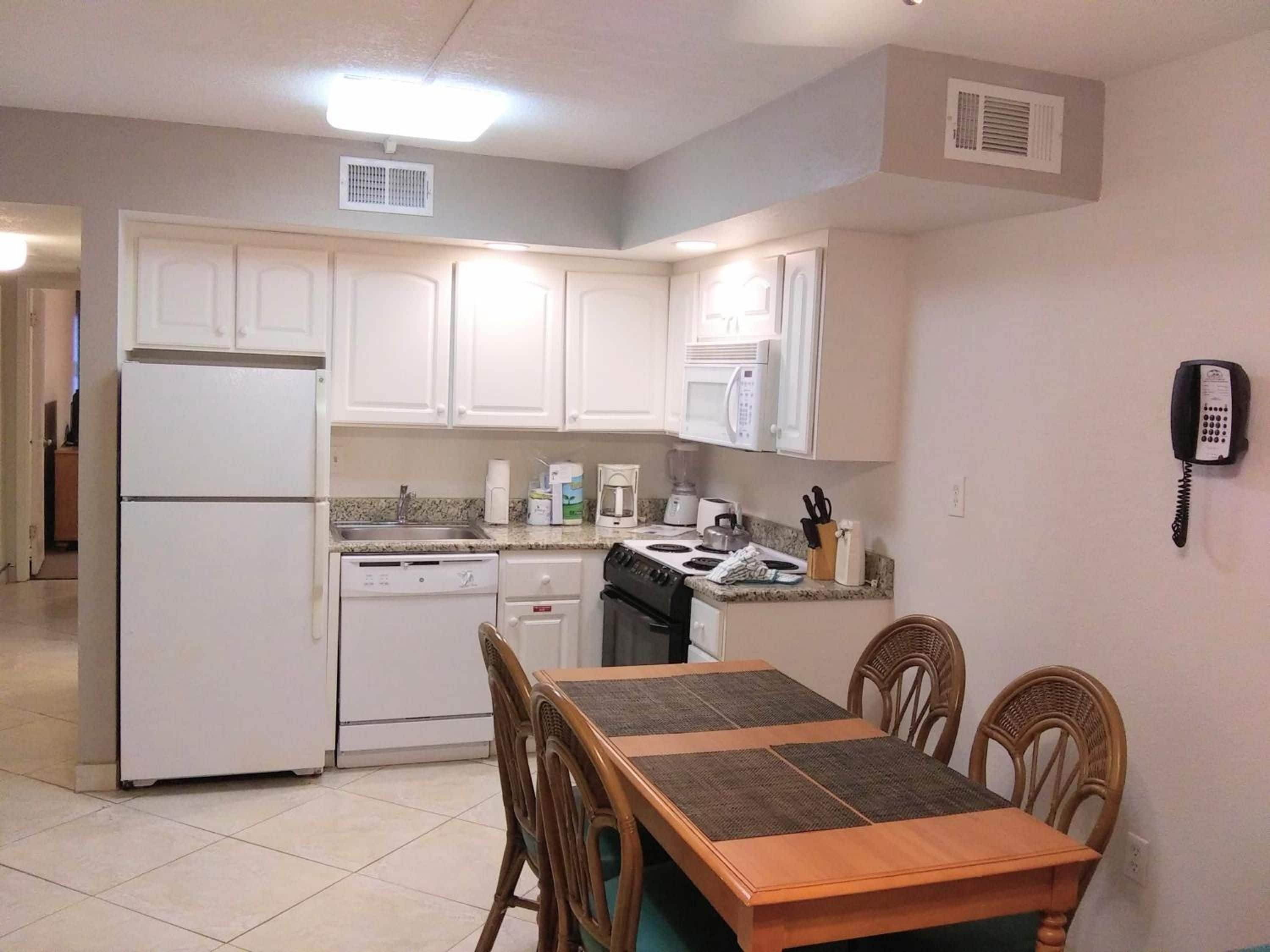 A compact kitchen with white cabinets, a refrigerator, a stove, and a microwave. There is a dining table with chairs and a phone on the wall.