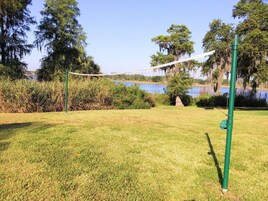 Een gazon met een volleybalnet, omringd door bomen en een waterpartij.