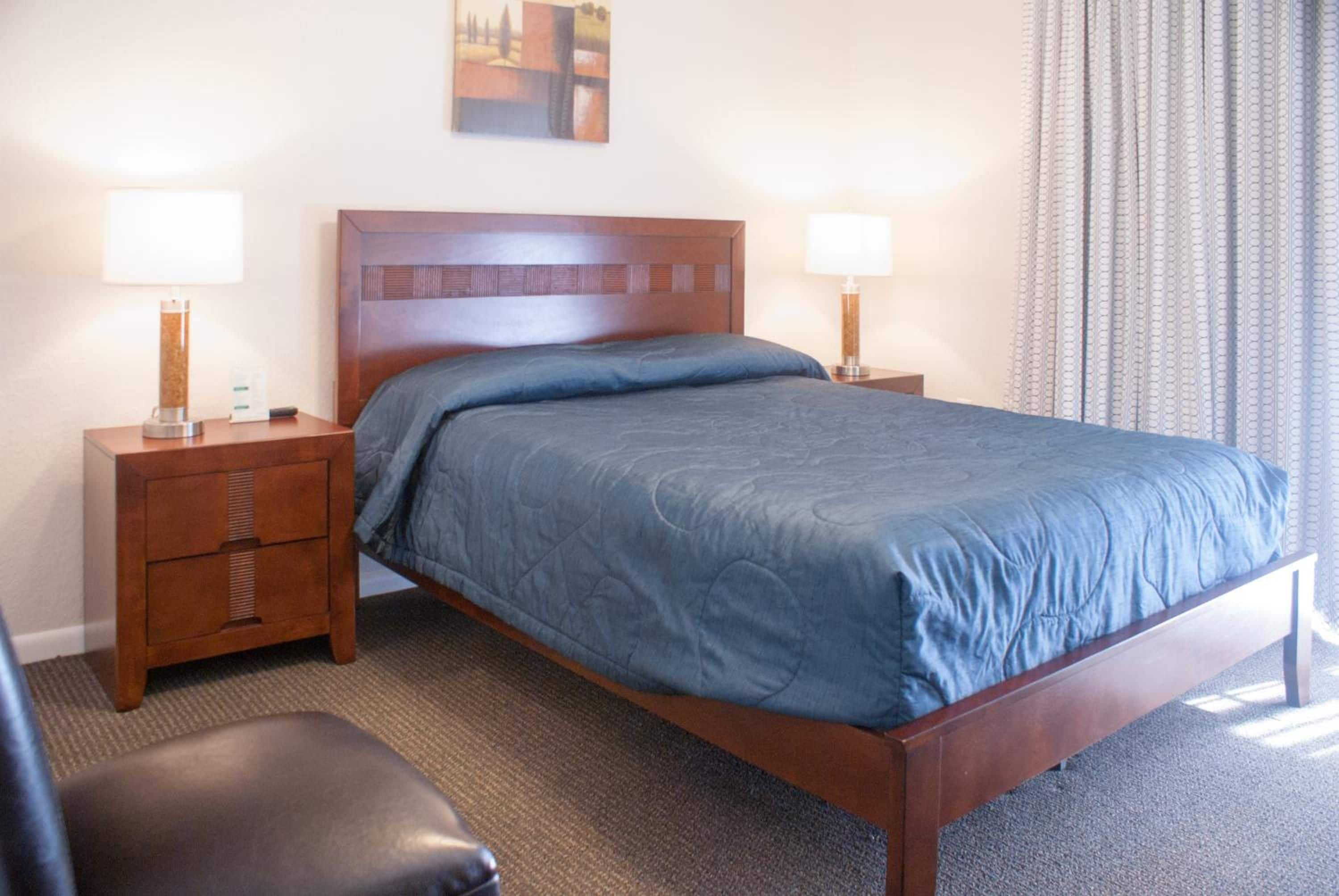A hotel room with a wooden bed, two bedside tables, a nightstand, a single chair, and a painting on the wall.