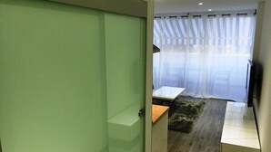 Una habitación de hotel moderna con una puerta corredera verde, una mesita de madera, un televisor montado en la pared y un ventanal con cortinas translúcidas.
