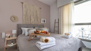 A neatly arranged bedroom with a bed, bedside tables, a wall clock, a macrame wall hanging, and a balcony view.