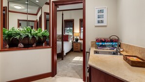 A hotel room with a large mirror, a bed with a red pillow, a nightstand with a lamp, a sink with a towel, and a window with blinds.
