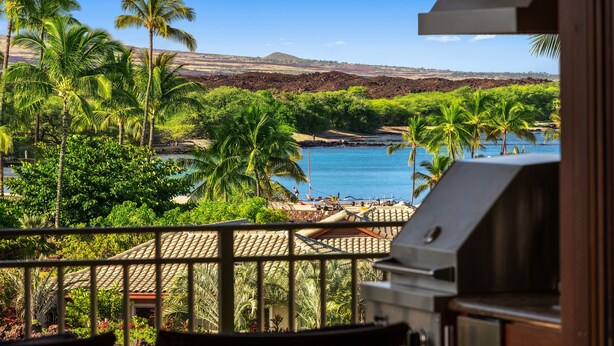 A balcony with a grill, overlooking a tropical landscape with palm trees and a view of a beach and water.