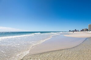 A beach with clear waters, a sandy shore, and a city skyline in the background.