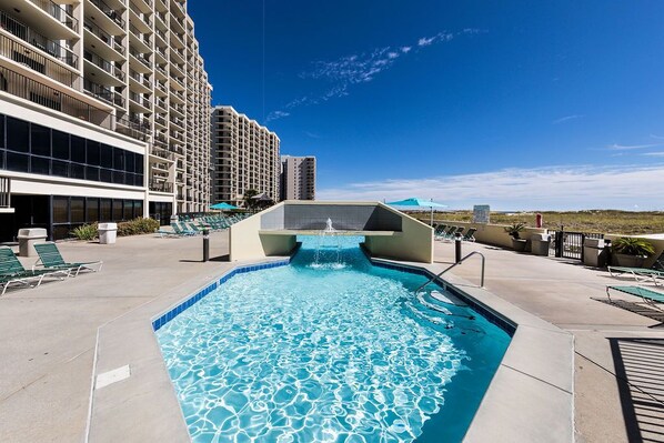 A swimming pool with a water feature, surrounded by lounge chairs and a hotel building.
