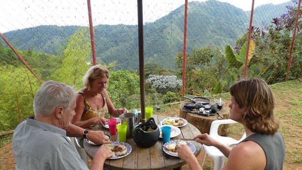 Un grupo de personas cenando en una mesa al aire libre con montañas al fondo.
