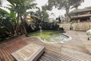 Una terraza de madera con un pequeño estanque, rodeada de plantas tropicales y una valla de madera.