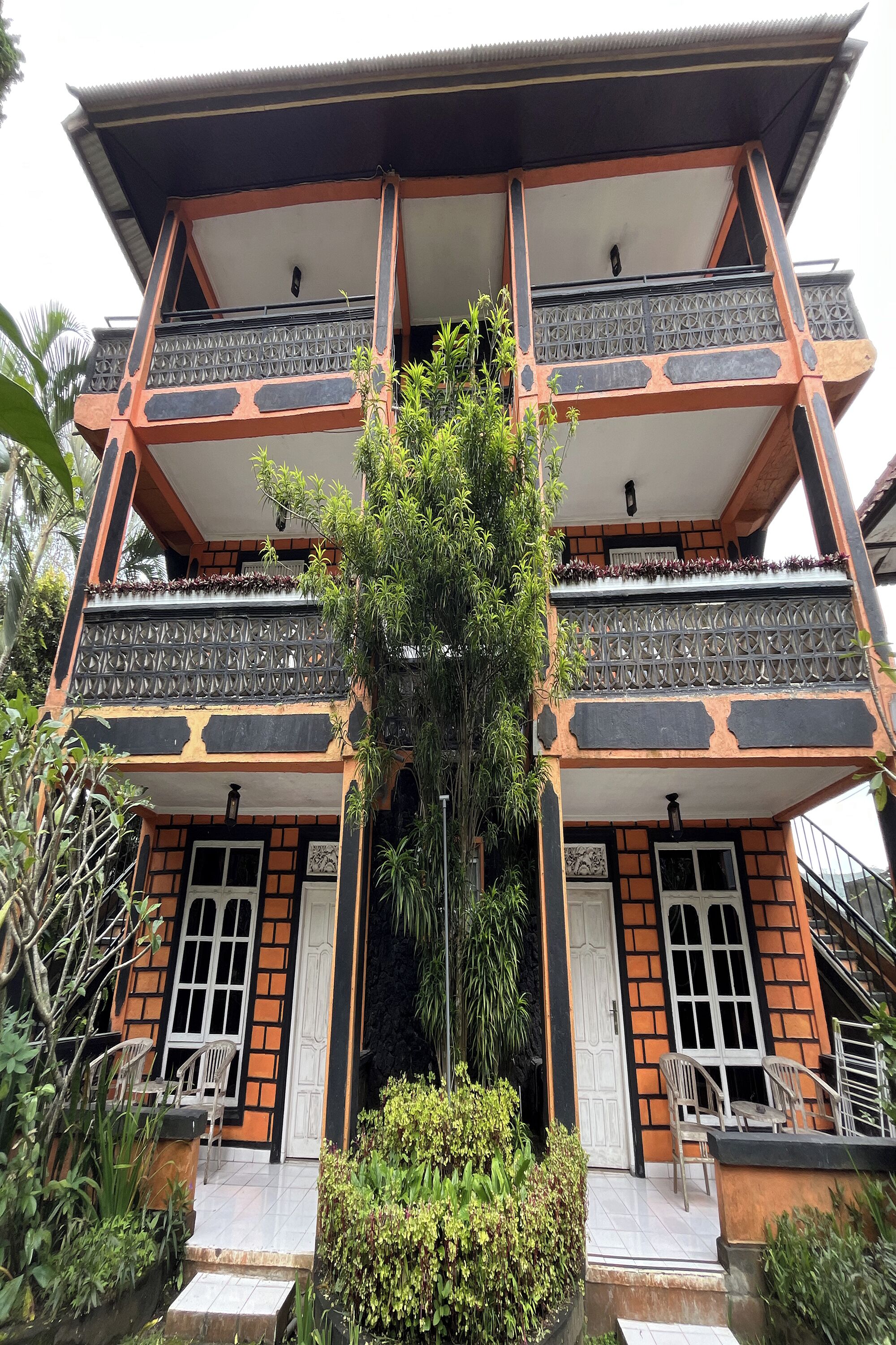 A multi-story building with a balcony on each floor, surrounded by greenery.