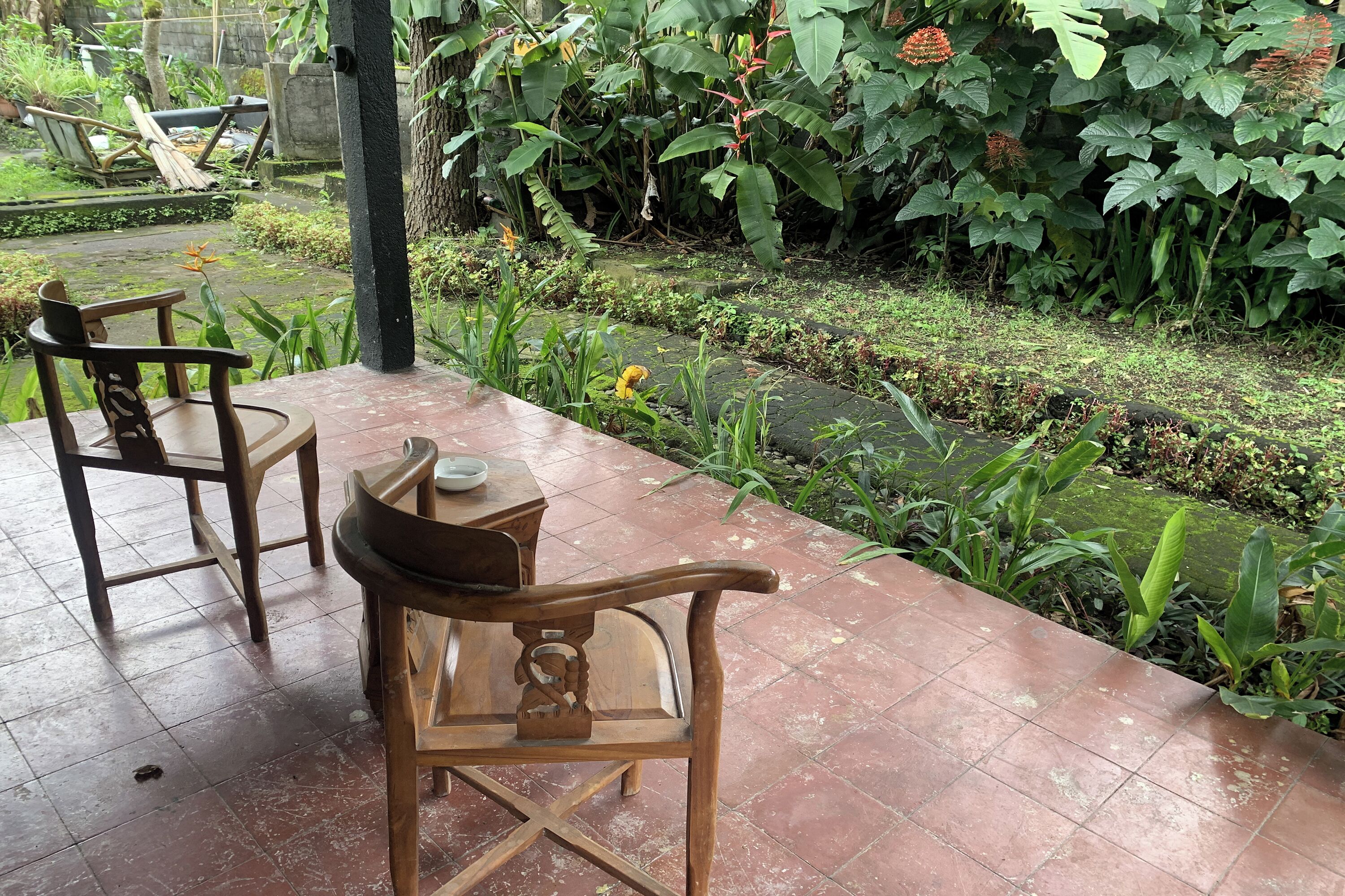 A patio area with two wooden chairs and a table, surrounded by lush greenery and a small water feature.