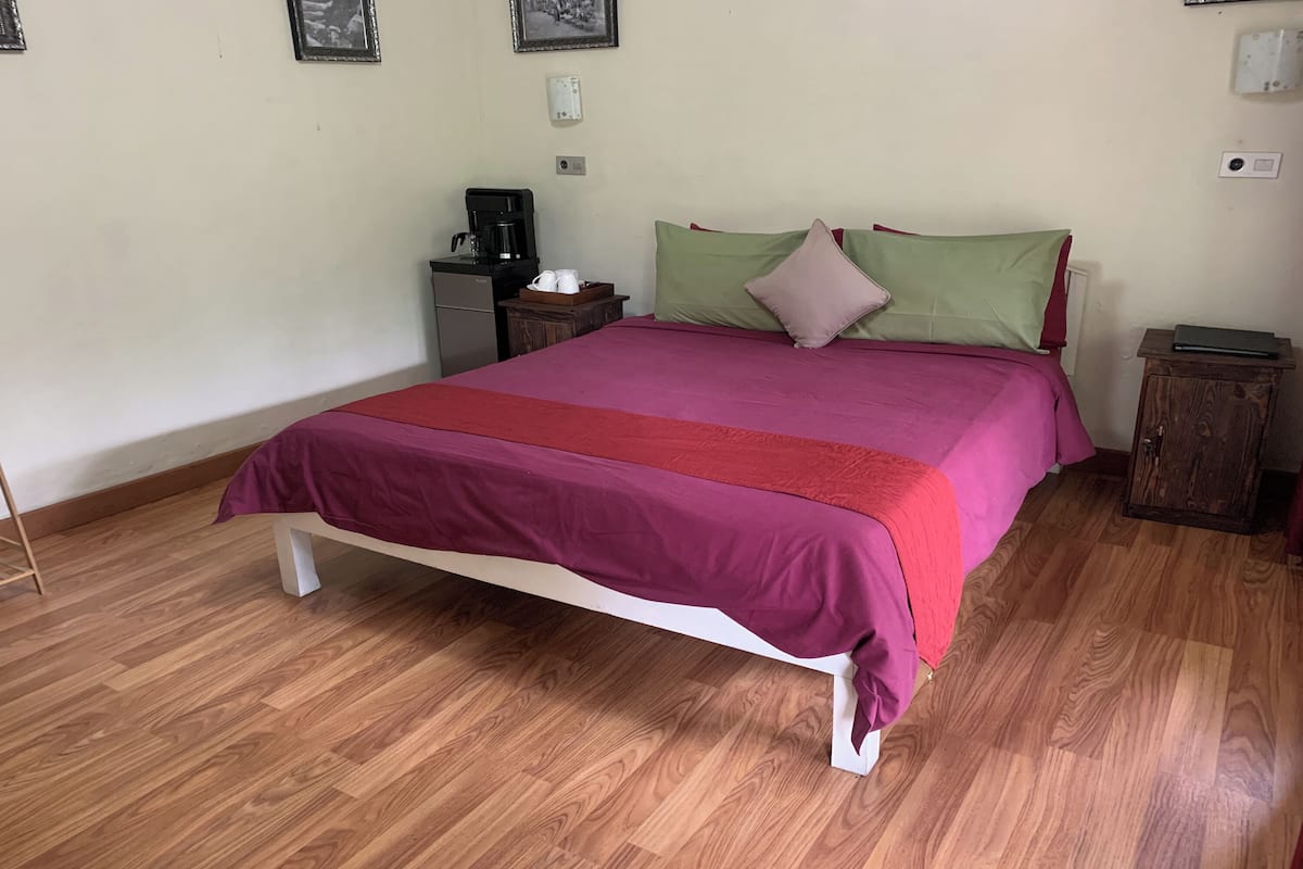 Una cama bien hecha con una colcha roja y rosa, una mesita de noche pequeña con una lámpara y una cafetera sobre una mesita auxiliar.