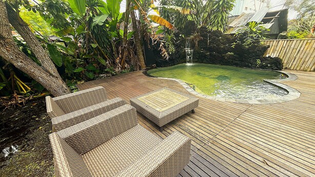 Une terrasse en bois avec des meubles en osier, un petit étang et des plantes tropicales.