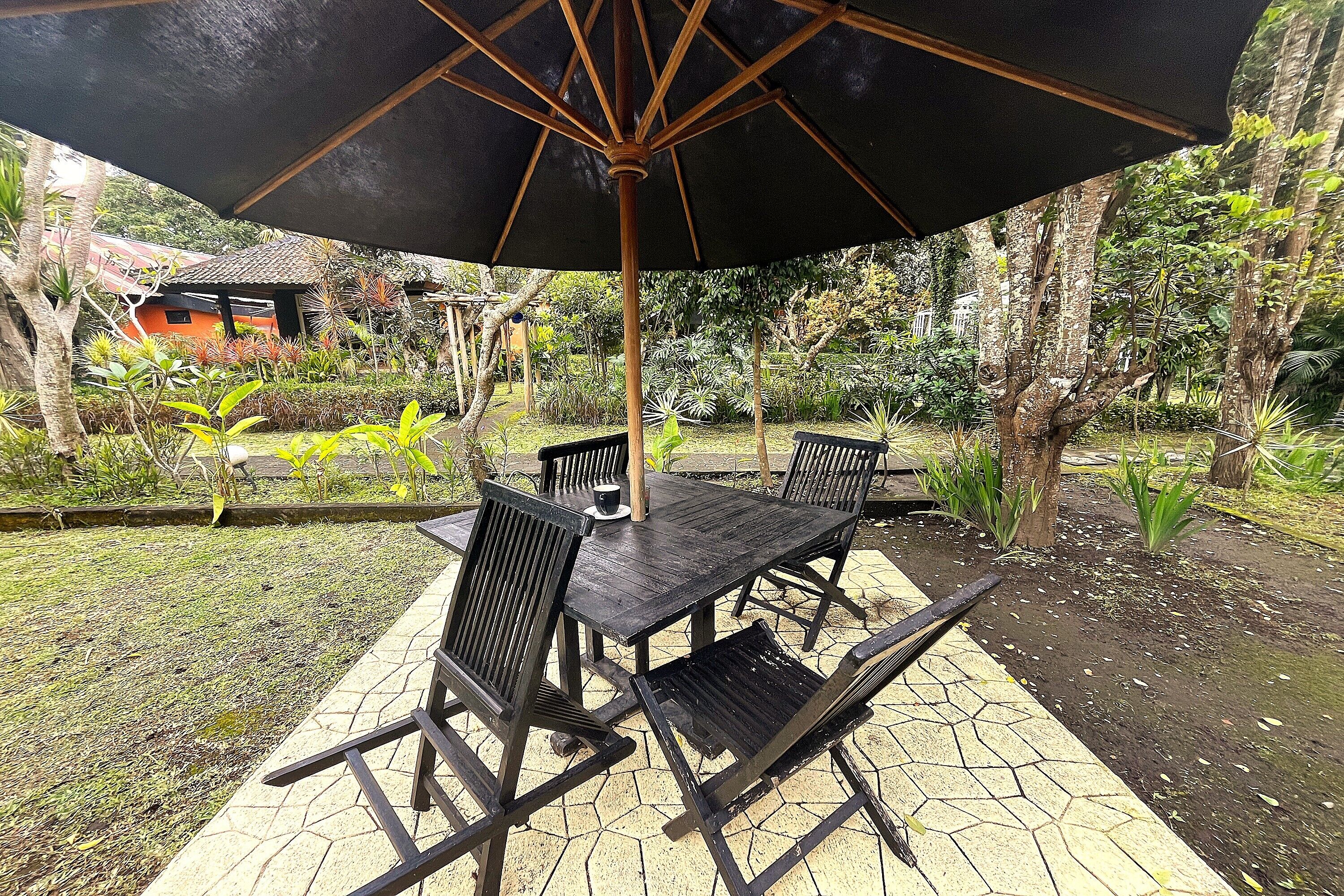 An outdoor patio with a table and chairs under a large umbrella, surrounded by lush greenery and a small building in the background.
