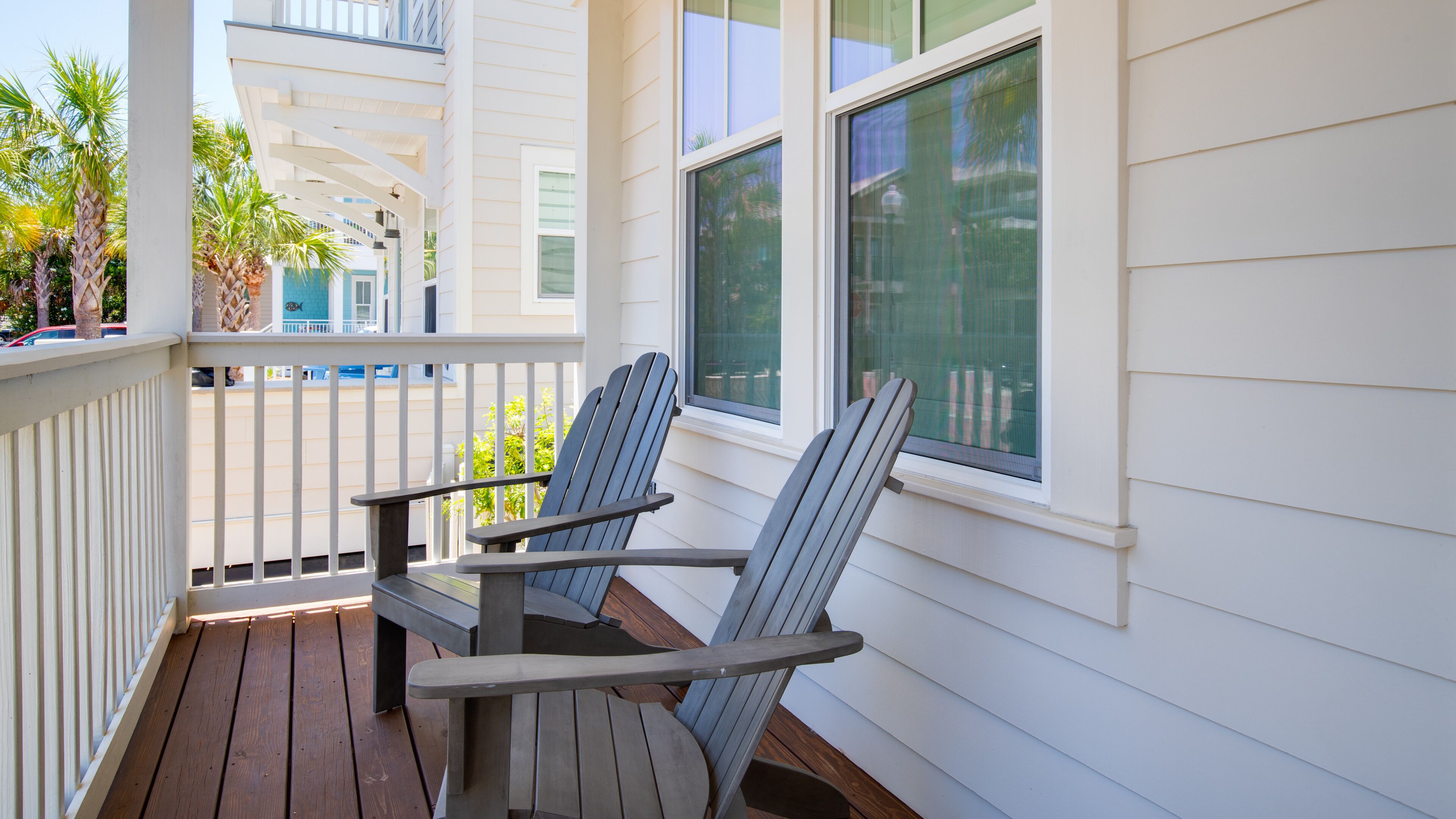 Una terraza de madera con sillas Adirondack, baranda blanca y vista a una calle con palmeras.