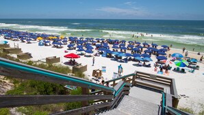 A beach with numerous umbrellas and sun loungers, a wooden boardwalk leading to the beach, and the ocean in the background.