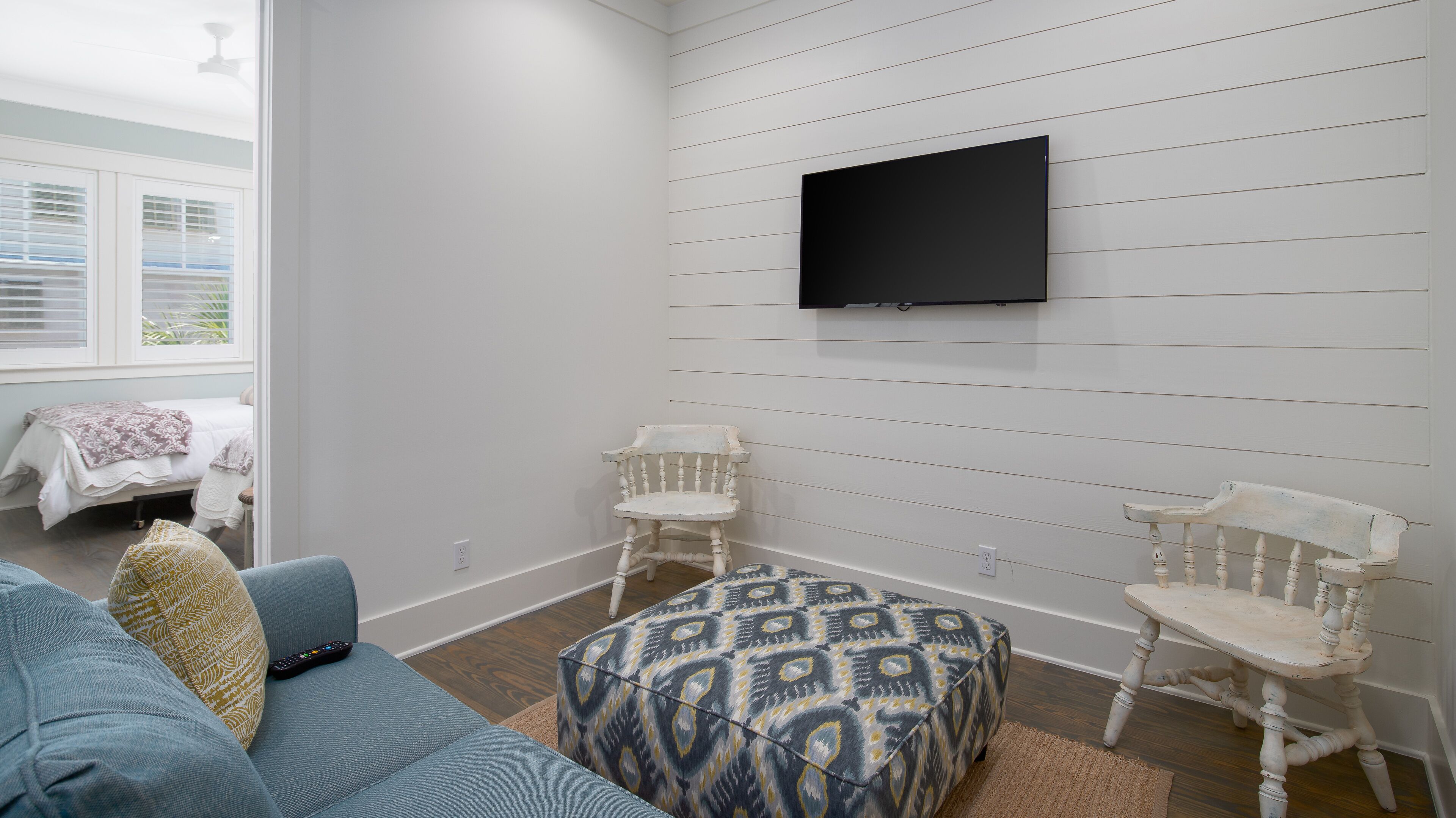 A room with a blue sofa, patterned ottoman, two white chairs, a TV mounted on the wall, and a bed with a white and brown blanket.