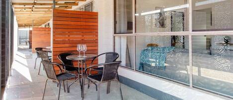 An outdoor seating area with a table and chairs, a wooden partition, and a view through large windows.