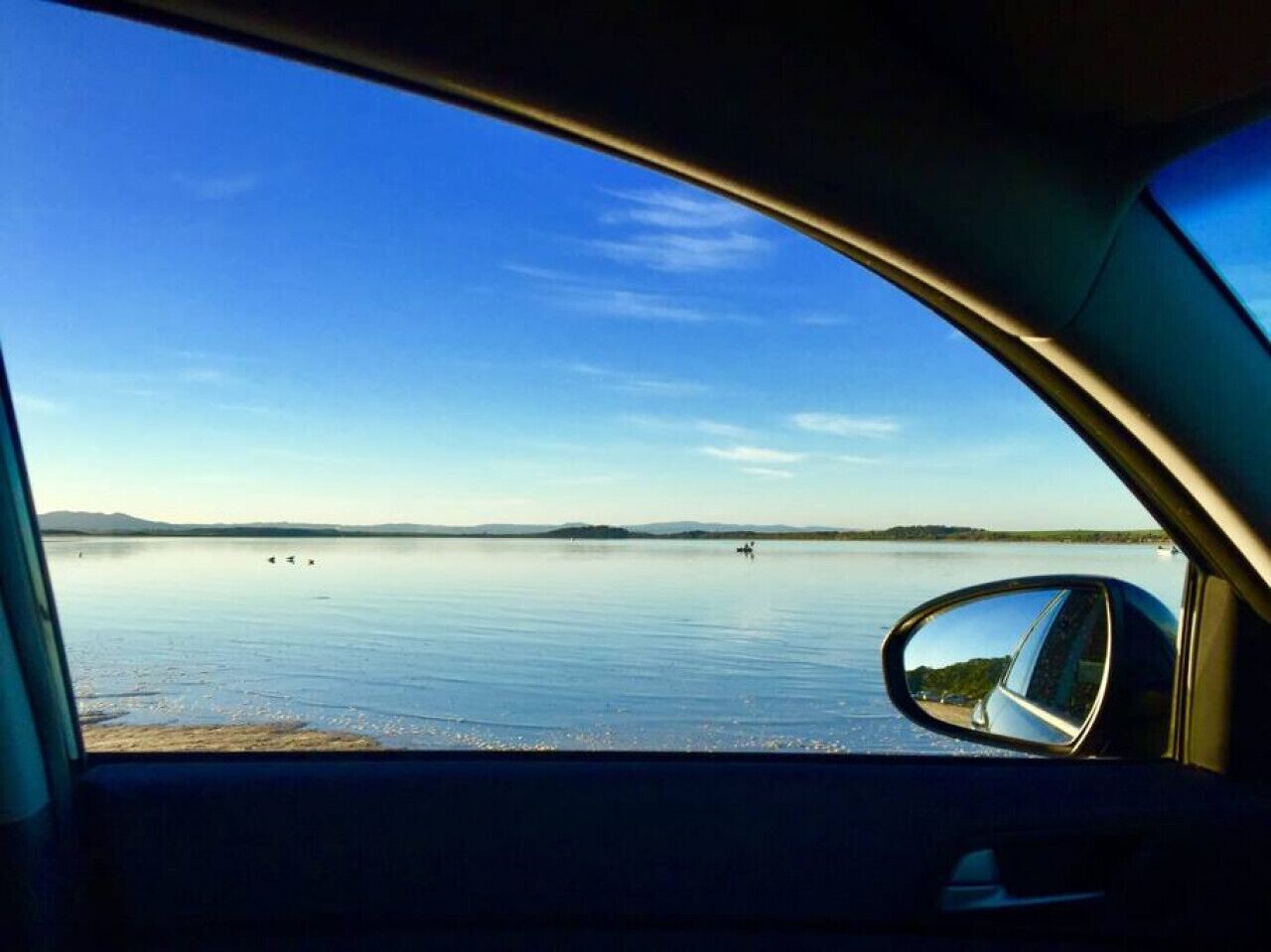 A car's side mirror reflecting a calm lake with a clear sky.