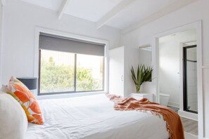 A bedroom with a large window, a bed with white bedding, a bedside table with a lamp, and a potted plant. - Prom Coast 1~Cosy beach life, stroll to the beach (Sandy Point)