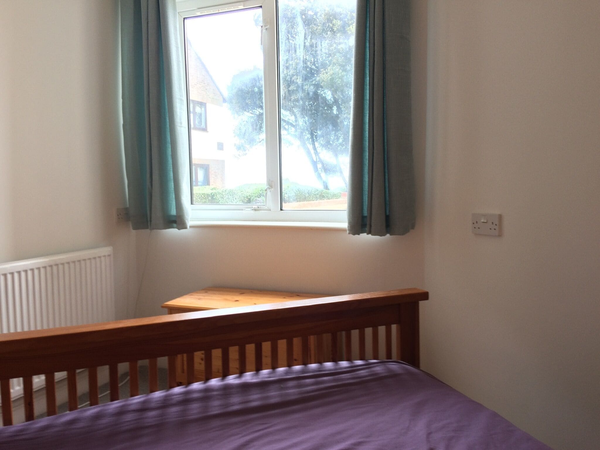 A bedroom with a wooden bed frame, a window with curtains, and a radiator.