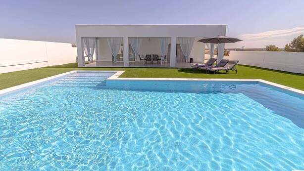 Una piscina rectangular con agua azul cristalina, rodeada por un edificio blanco con un porche cubierto.