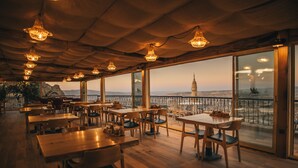 A restaurant with wooden tables and chairs, large windows with a view of a distant tower, and hanging pendant lights. - Hu of Cappadocia (Nevsehir)