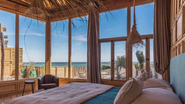 A bedroom with a large bed, a chair, and a view of the beach and ocean through large windows. - Palais Rhoul Dakhla (Dakhla)