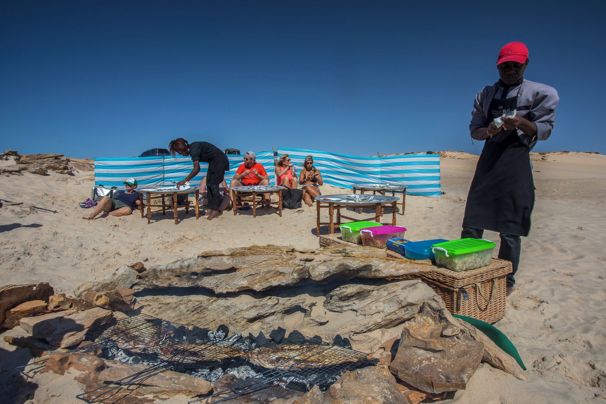 A beach scene with people sitting on chairs under a striped canopy, a grill set up on rocks, and a person standing with a bag.