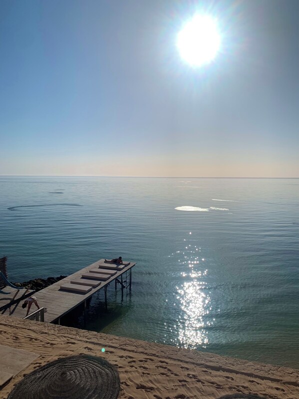 A beach with a wooden pier extending into calm waters, a sandy area with footprints, and a clear sky.