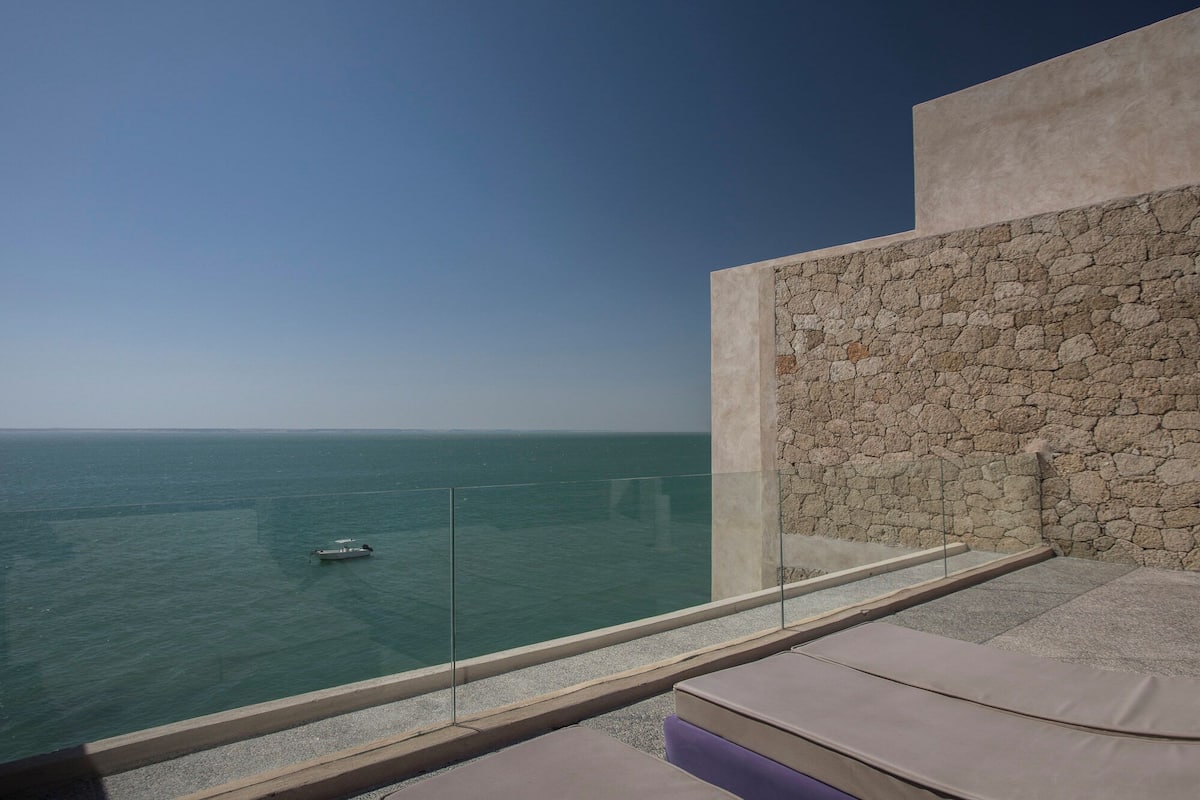 A balcony with a view of the sea, a stone wall, and a small boat in the distance.