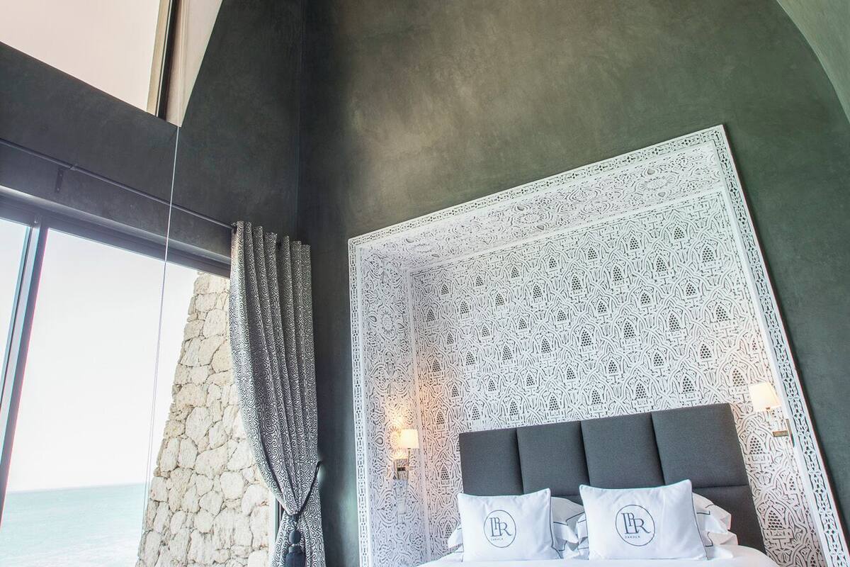 A modern bedroom with a large bed, a patterned headboard, and a view of the sea.