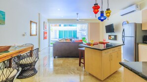 Una cocina moderna con una isla central, una zona de comedor con una mesa y sillas de madera, y un espacio de sala de estar con sofá y vistas al exterior.