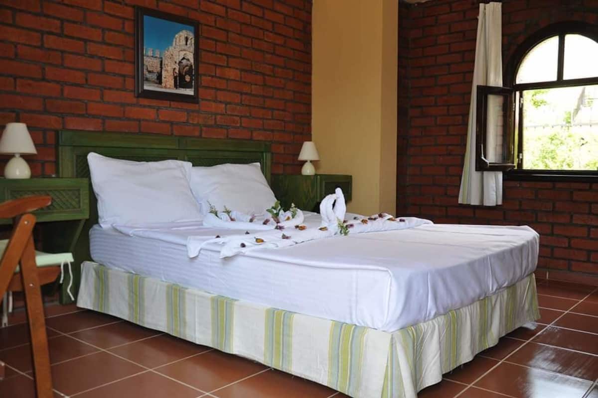 Una cama doble con sábanas blancas y adornos florales, cabecera verde y pared de ladrillo con un cuadro enmarcado.