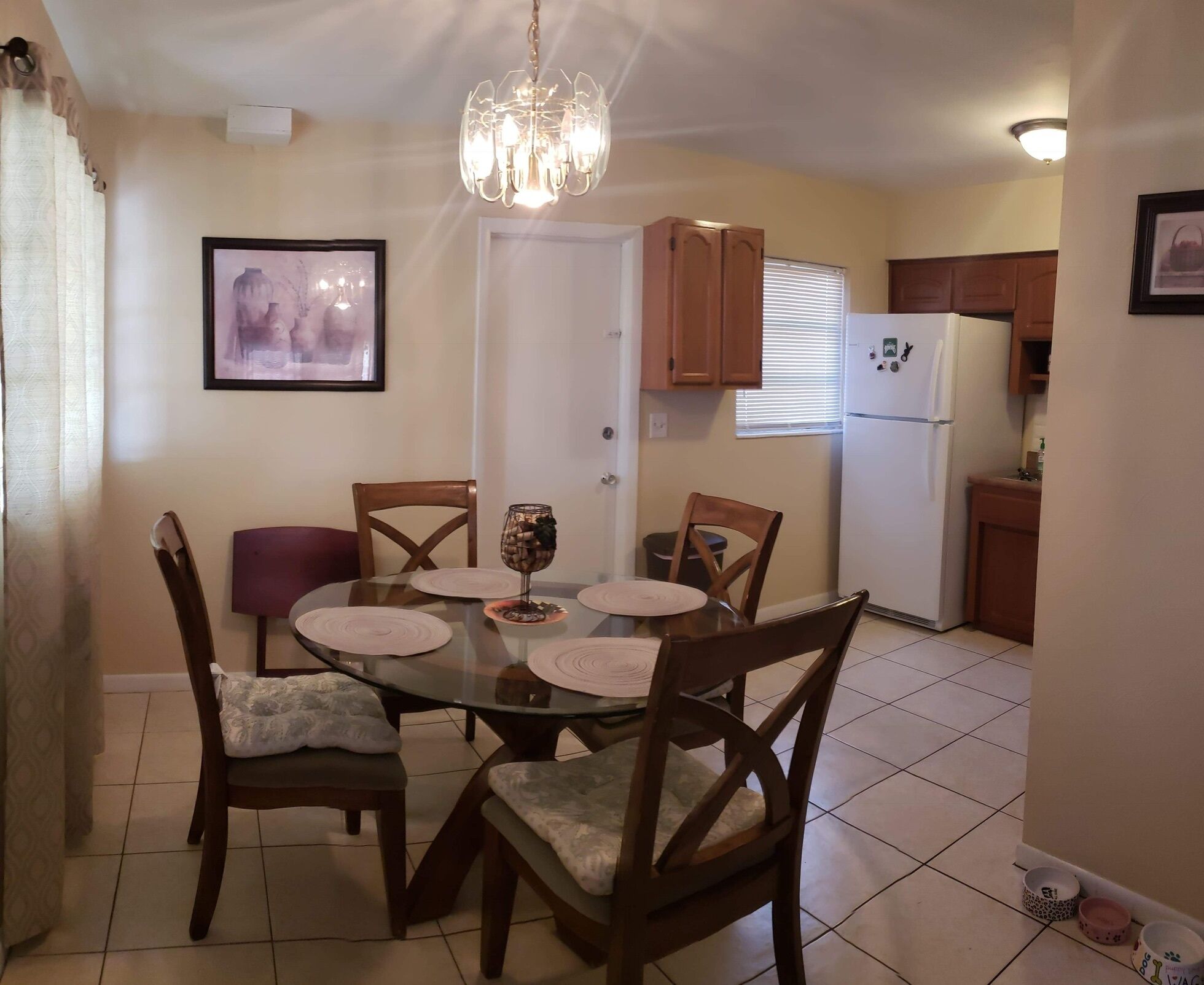 A dining area with a glass table surrounded by wooden chairs, a chandelier, and a framed picture on the wall.