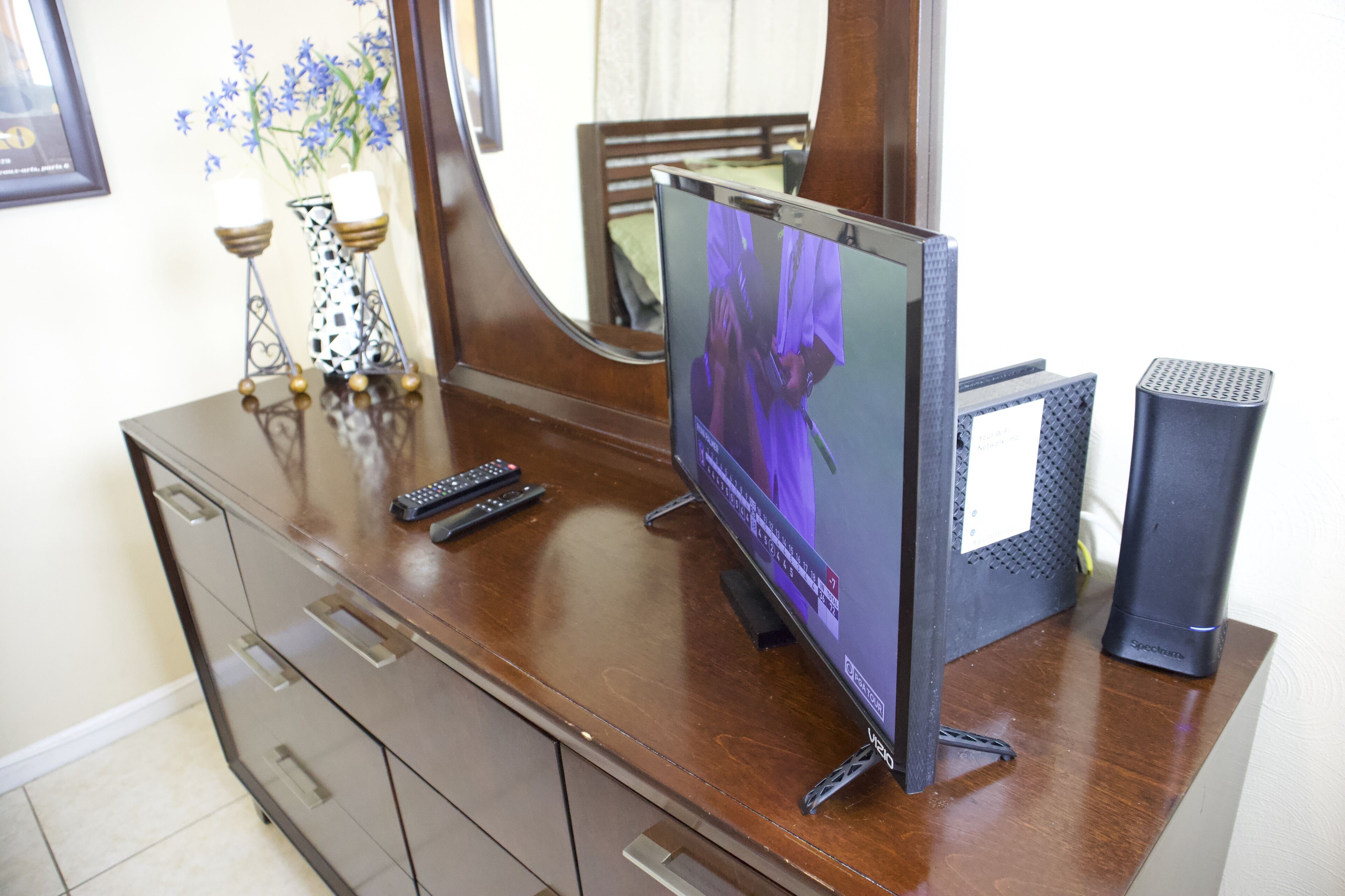 Un televisor de pantalla plana sobre un aparador de madera con un espejo encima, un altavoz pequeño y un florero con flores.
