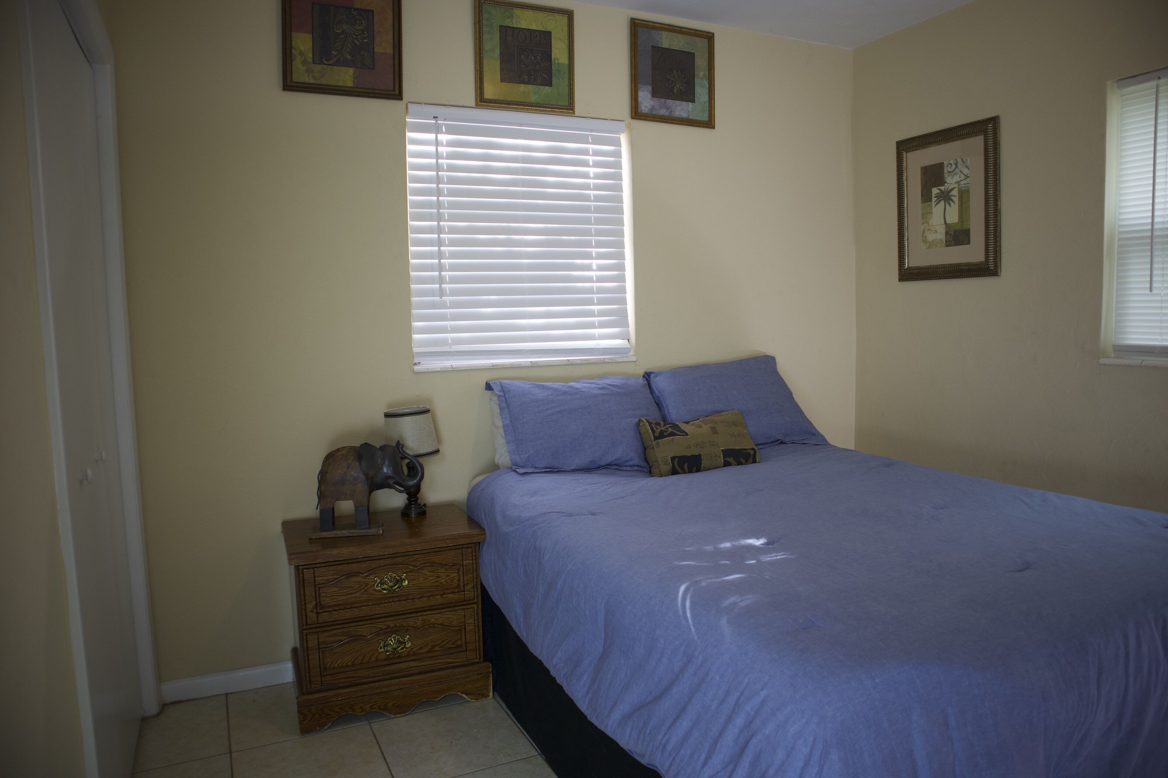 Un dormitorio con cama, una mesita de noche, una ventana con persianas y tres cuadros enmarcados en la pared.