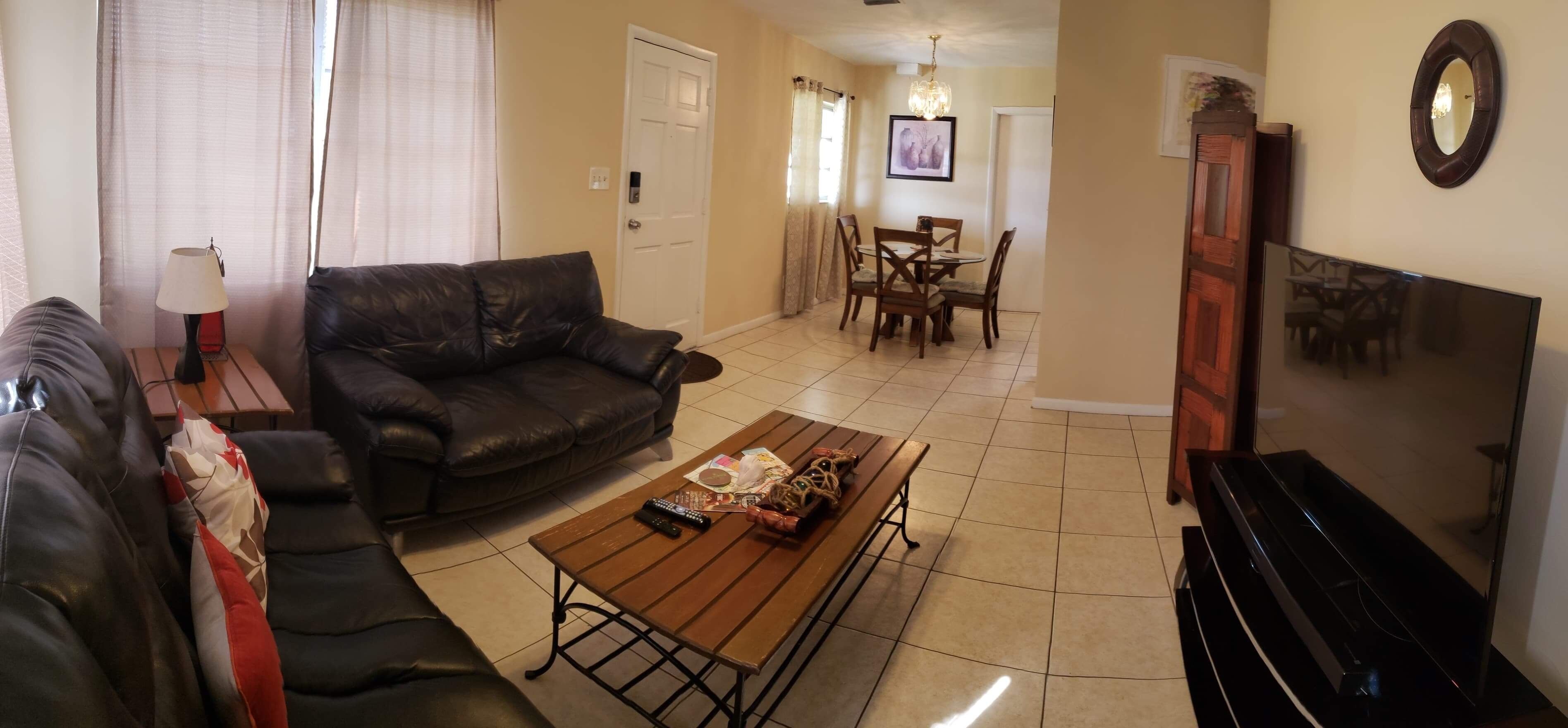 Una sala de estar con un sofá de cuero negro, una mesa de centro de madera con controles remotos, un comedor con una mesa y sillas de madera, y un mueble de televisión negro.