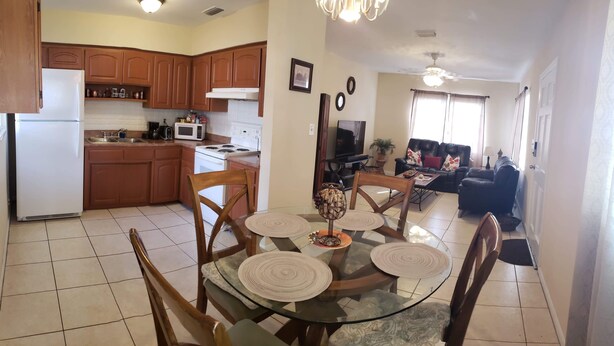 Cocina compacta con armarios de madera, un refrigerador, un microondas y una estufa. Hay un comedor con mesa y sillas de vidrio, y una sala de estar con sofá y televisión.