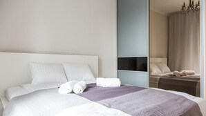A neatly made bed with white and gray bedding, a bedside table with a lamp, and a mirror reflecting part of the room.