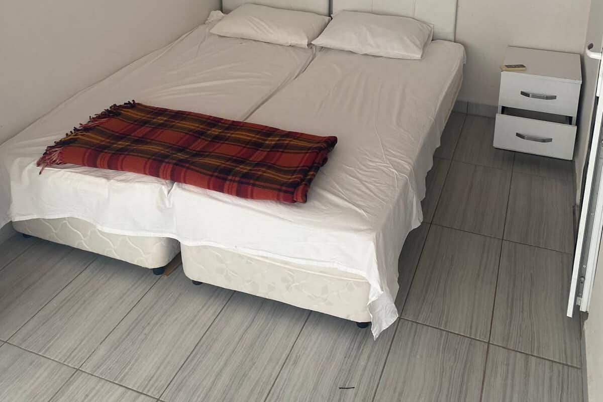 A single-bed room with a white headboard, a bedside table with drawers, and a red plaid blanket on the bed.