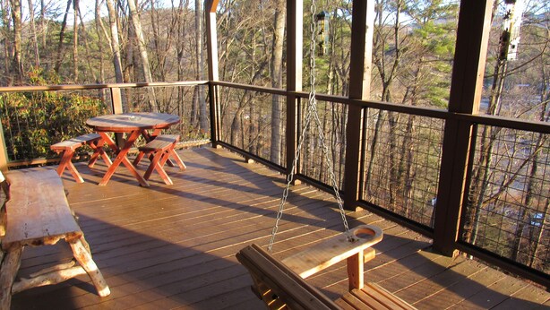 A wooden deck with picnic tables and benches, surrounded by a metal railing, overlooking a forested area.