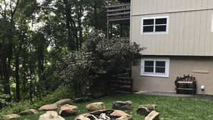 A backyard with a fire pit surrounded by rocks, a wooden deck, and a house with multiple windows.