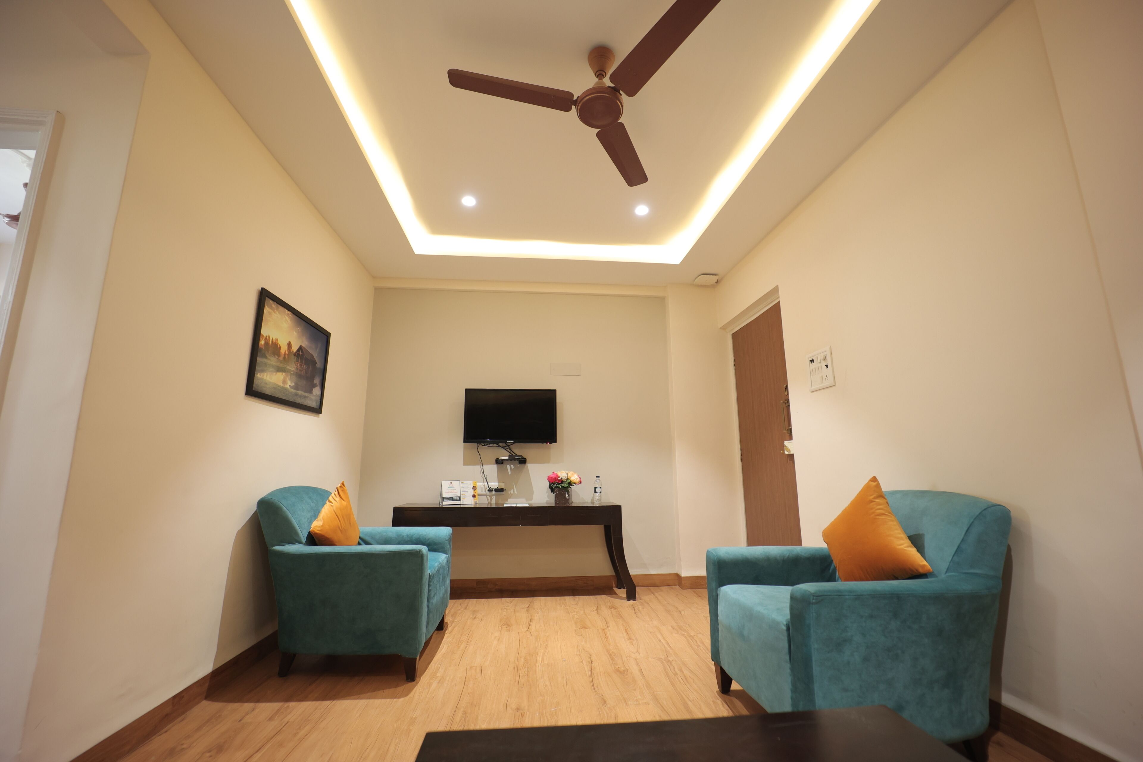 A modern living room with teal armchairs, a dark wooden coffee table, a flat-screen TV, and a ceiling fan.