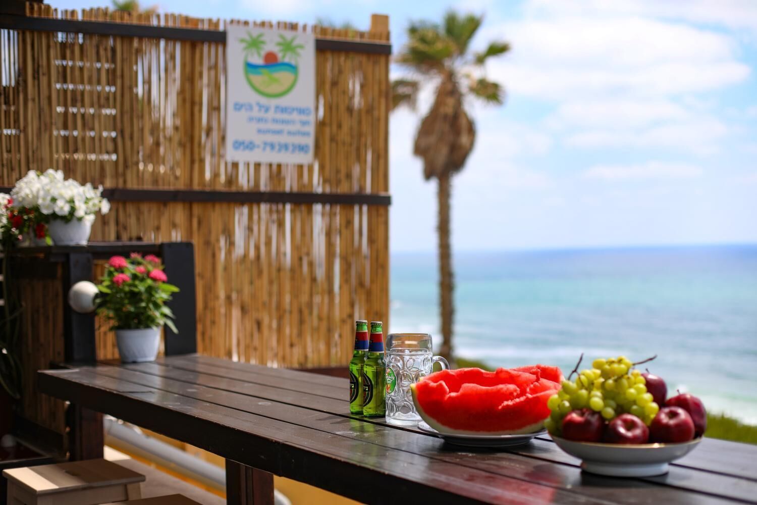 Une table en bois sur laquelle se trouvent un saladier de fruits, un verre d’eau et des bouteilles de bière, le tout placé devant une clôture en bois et avec une vue sur l’océan.