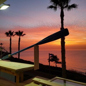 A sunset view from a wooden deck with a metal railing, overlooking the ocean and palm trees.