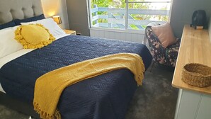 Ein Schlafzimmer mit Bett, Nachttisch, Fenster mit Blick auf Grünflächen und einer kleinen Holzablage.