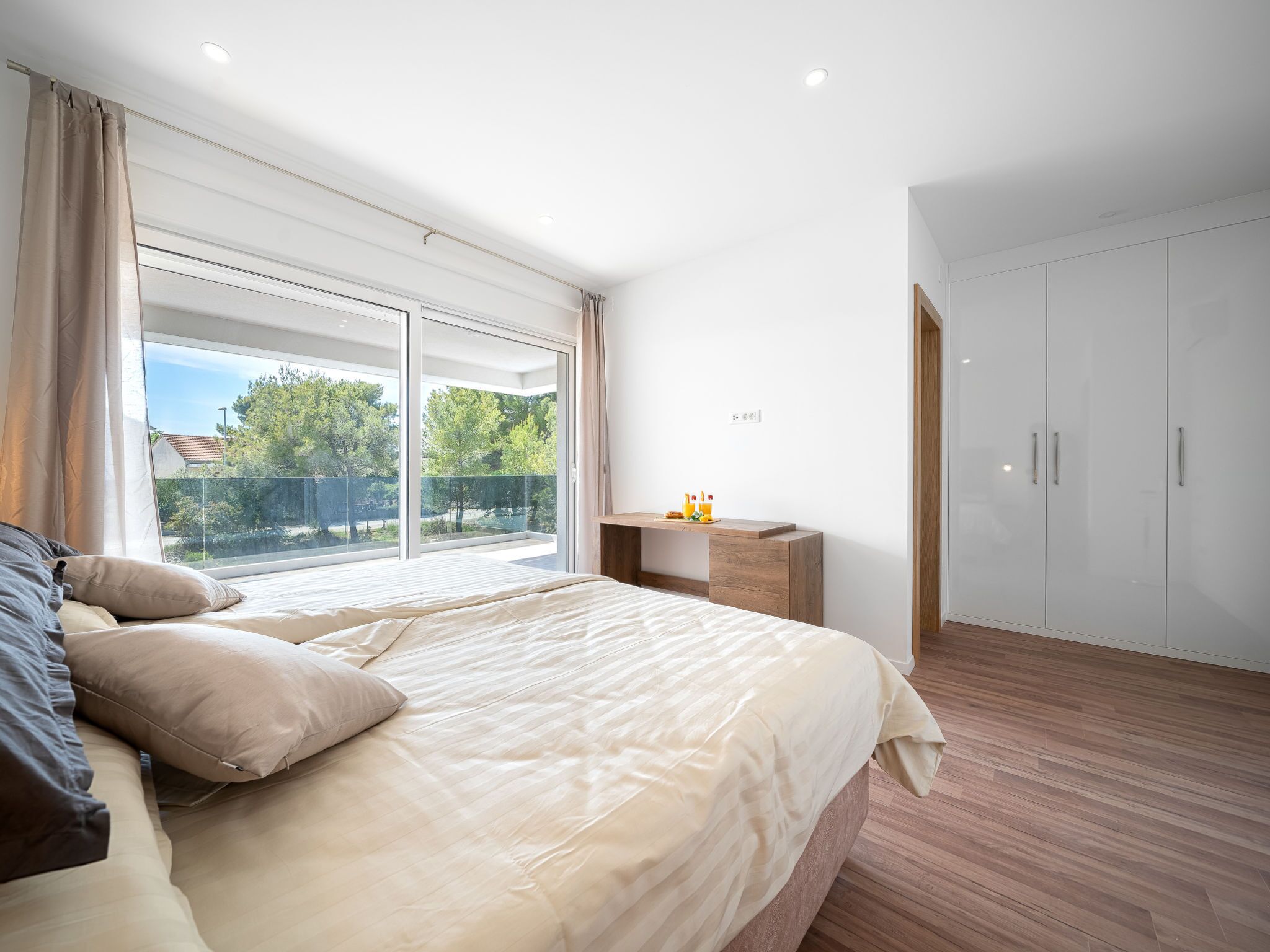 A modern bedroom with a large bed, a wooden nightstand, a wardrobe, and a sliding glass door to a balcony.