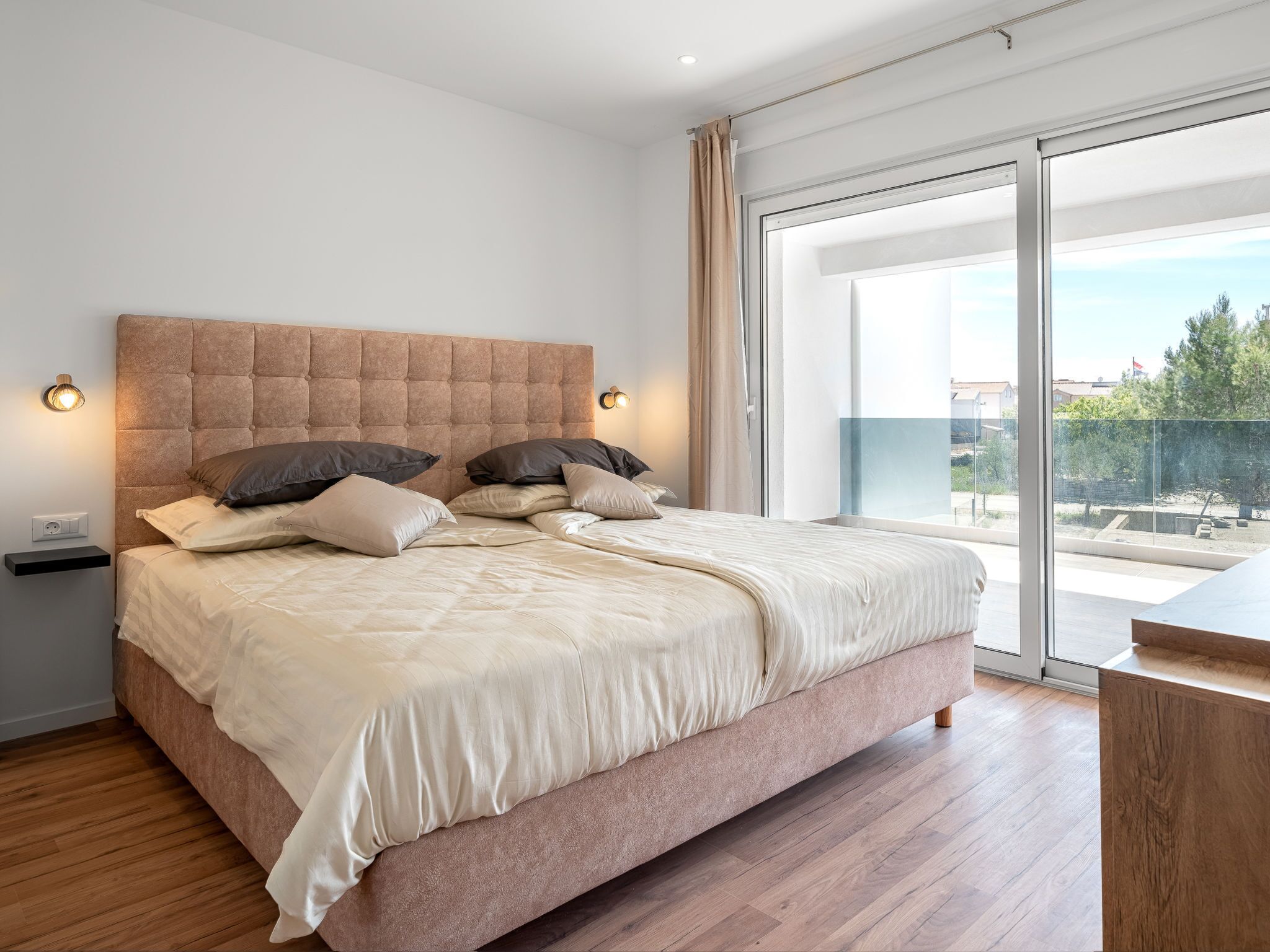 A modern bedroom with a large bed, a wooden floor, and a view of the outdoors through a sliding glass door.