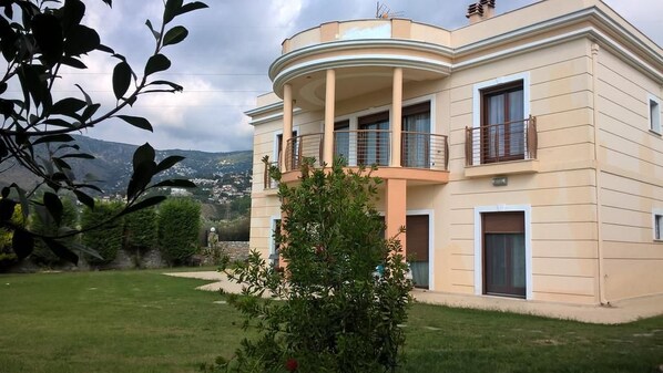 A two-story building with a curved balcony, a small garden, and a view of the surrounding landscape. - Luxury Villa Thea (Volos)